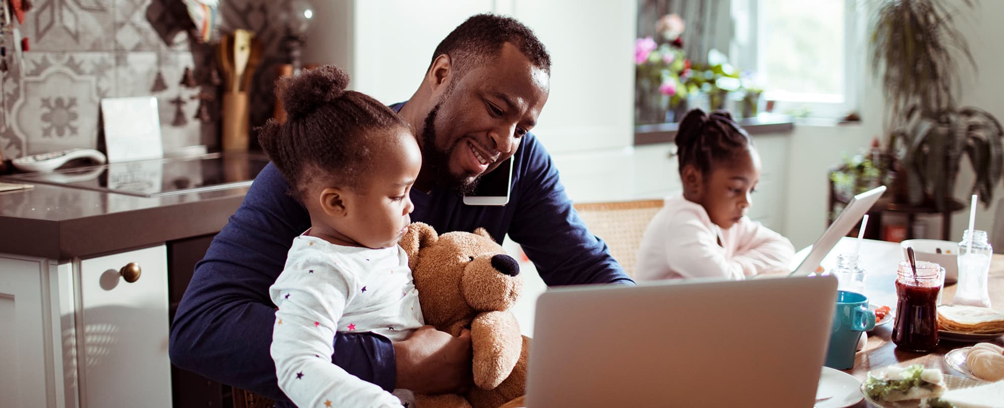 Man with his two daughters at the kitchen table, man is holding one of his daughters and her teddy bear as he talks on the phone and works on his laptop