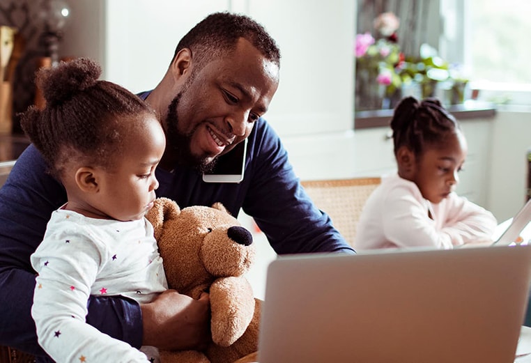 Man with his two daughters at the kitchen table, man is holding one of his daughters and her teddy bear as he talks on the phone and works on his laptop