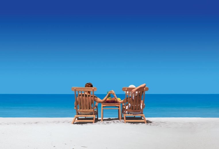 Couple on the beach facing the ocean sitting in beach lounge chairs holding hands over a side table 