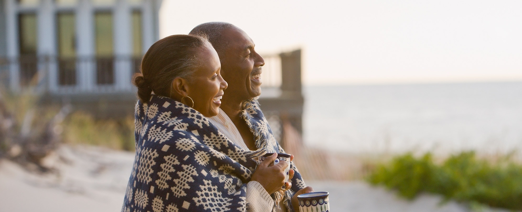 An older couple wrapped up in a blanket smile, looking out over the ocean.